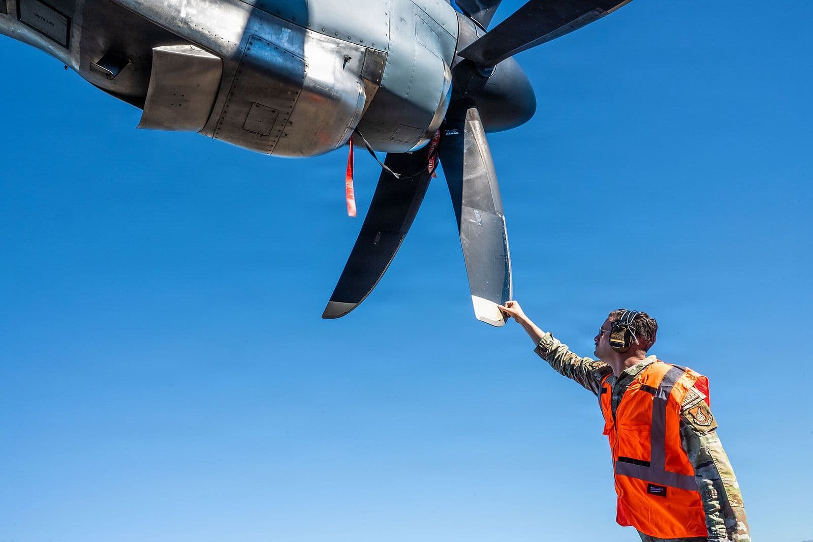 A technician in a high-visibility vest inspects a turboprop propeller blade against a clear blue sky